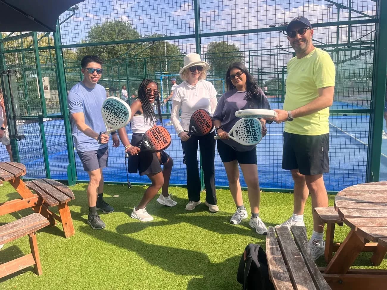 Five people holding padel rackets beside a court, laughing